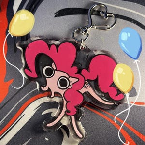 May include: A clear acrylic keyring featuring a pink cartoon pony with a heart-shaped tail and a heart-shaped clasp. The pony is holding two yellow balloons and a blue balloon. The background has a grey and red pattern.