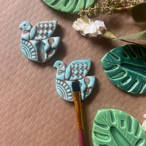 May include: A collection of ceramic art pieces, including two turquoise bird-shaped ornaments with brown detailing and green leaf-shaped dishes. A paintbrush with a red handle rests on one of the bird ornaments. White flowers and greenery are also present.
