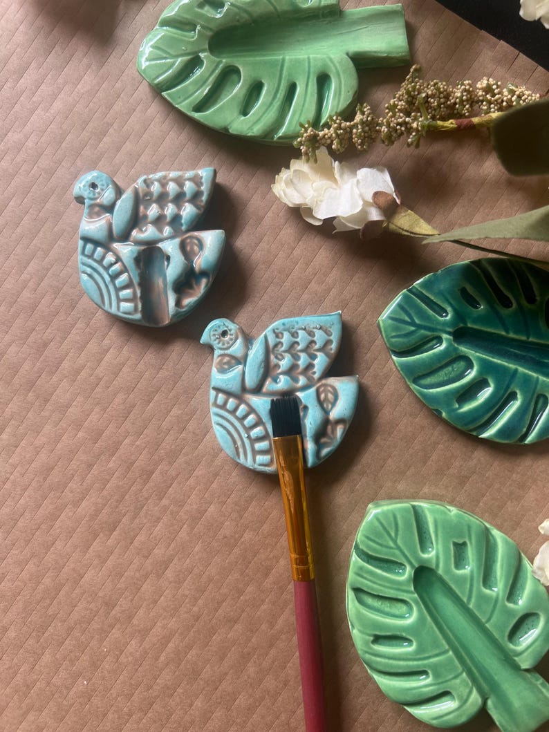 May include: A collection of ceramic art pieces, including two turquoise bird-shaped ornaments with brown detailing and green leaf-shaped dishes. A paintbrush with a red handle rests on one of the bird ornaments. White flowers and greenery are also present.