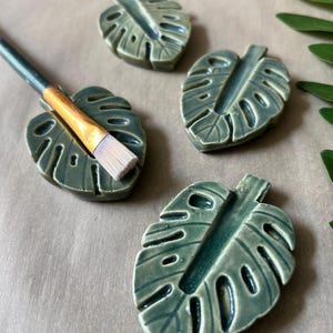 May include: Four ceramic leaf-shaped paint brush holders in shades of green. One holder has a paint brush resting in it. The holders are arranged on a neutral-toned surface, with a few green leaves in the background.