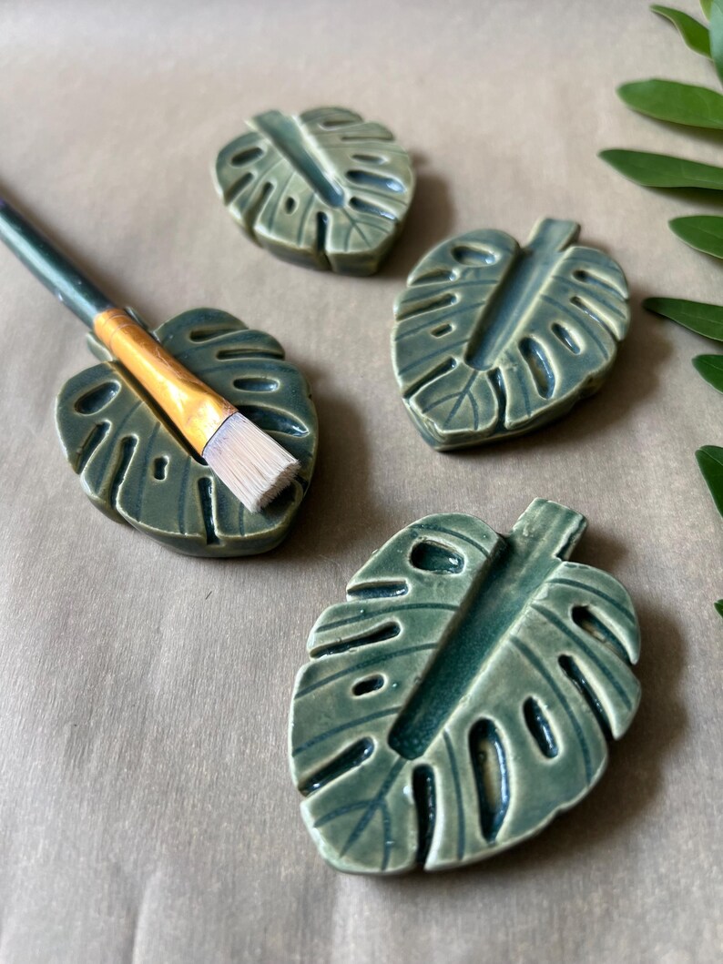 May include: Four ceramic leaf-shaped paint brush holders in shades of green. One holder has a paint brush resting in it. The holders are arranged on a neutral-toned surface, with a few green leaves in the background.