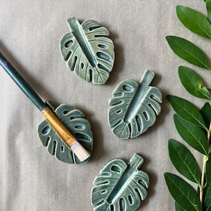 May include: Four ceramic leaf-shaped paint brush holders in a muted teal color. One holder has a paint brush resting in it. The holders are arranged on a neutral-toned surface, with green leaves in the background.