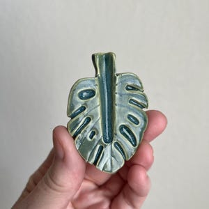 May include: A ceramic leaf-shaped decorative item with a glossy finish. The leaf is primarily light green with darker green accents in the veins and edges. The design mimics a Monstera plant leaf.