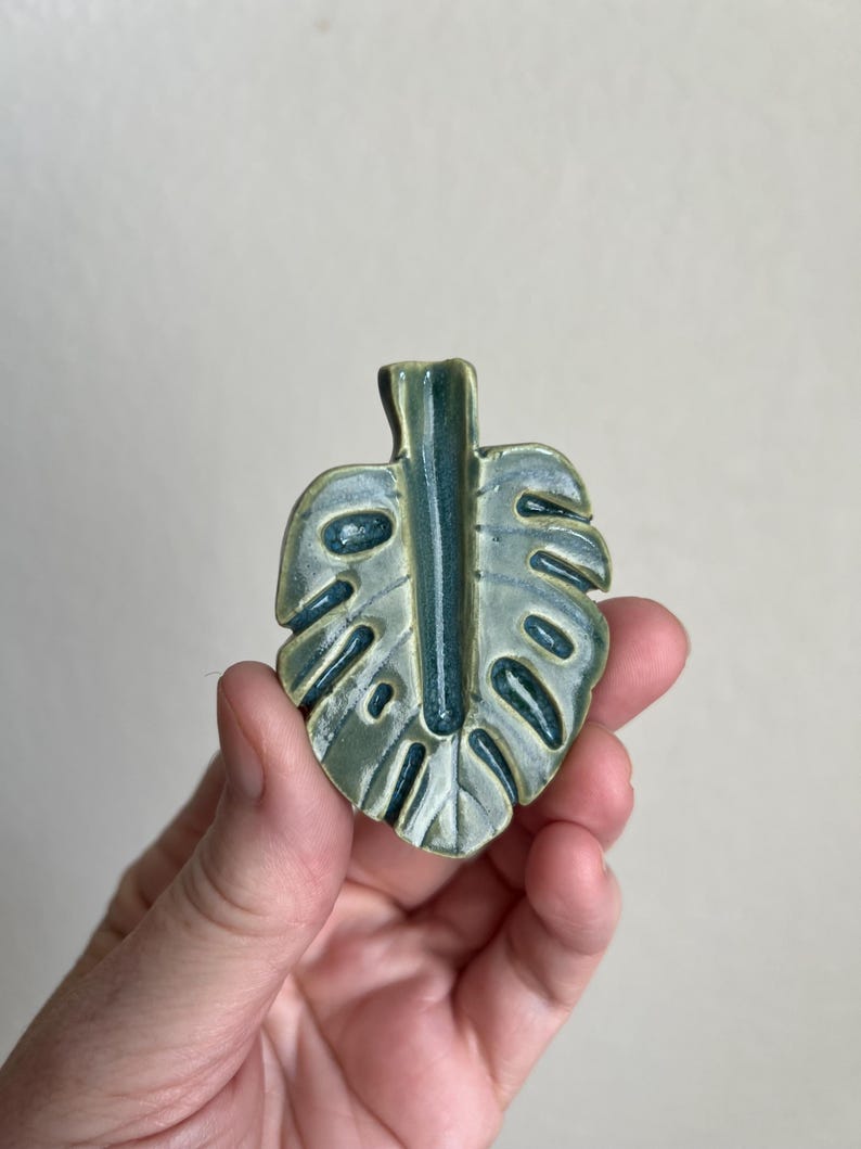 May include: A ceramic leaf-shaped decorative item with a glossy finish. The leaf is primarily light green with darker green accents in the veins and edges. The design mimics a Monstera plant leaf.