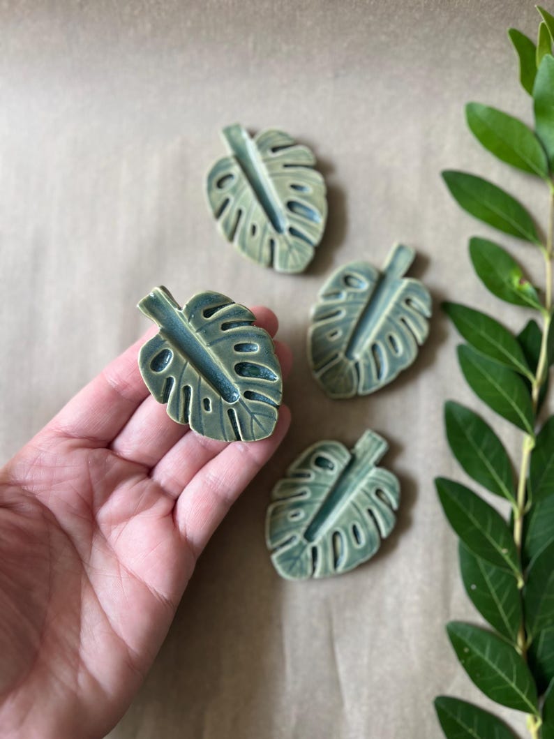 May include: Four ceramic monstera leaf-shaped objects in shades of green and blue. The leaves have detailed veining and are arranged on a neutral background. A sprig of green leaves is in the background.