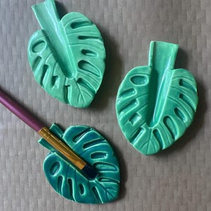 May include: Three green ceramic monstera leaf-shaped holders. One holder has a makeup brush resting in it. The holders are a bright, glossy green color and have detailed leaf vein patterns. The background is a neutral brown.