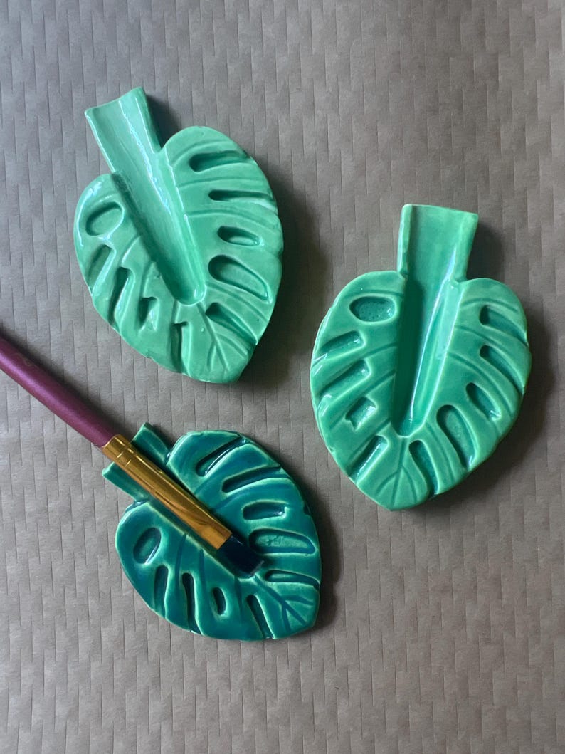 May include: Three green ceramic monstera leaf-shaped holders. One holder has a makeup brush resting in it. The holders are a bright, glossy green color and have detailed leaf vein patterns. The background is a neutral brown.