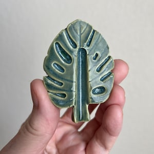 May include: A ceramic leaf-shaped decorative item in shades of green and blue. The leaf has a glossy finish and a detailed vein pattern. The item is held in a hand against a neutral background. The leaf measures approximately 3 inches in length.