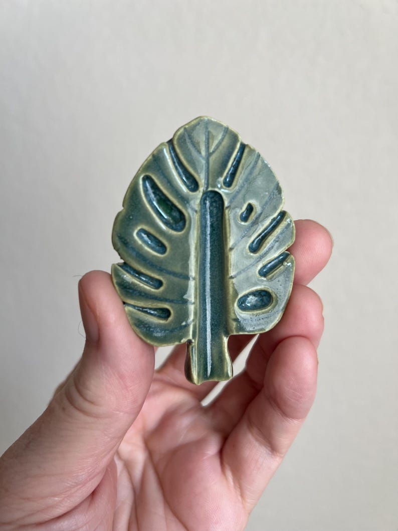 May include: A ceramic leaf-shaped decorative item in shades of green and blue. The leaf has a glossy finish and a detailed vein pattern. The item is held in a hand against a neutral background. The leaf measures approximately 3 inches in length.