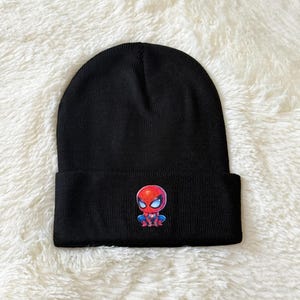May include: Black beanie featuring a colorful Spider-Man graphic. The graphic depicts a cartoon version of Spider-Man in red, blue, and white. The beanie is made of a soft, knitted material.