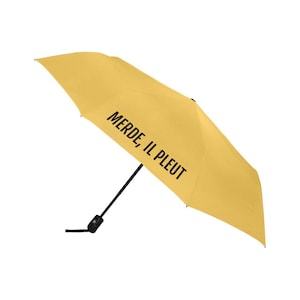 Peut inclure: Un parapluie jaune moutarde ouvert, avec l'inscription noire "MERDE, IL PLEUT". La structure et la poignée du parapluie sont noires.