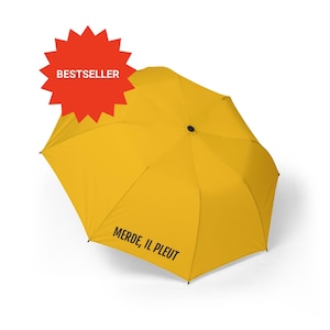 May include: Bright yellow umbrella with the text "MERDE, IL PLEUT" printed on the canopy. A red starburst with the word "BESTSELLER" is in the upper left corner. The umbrella is open and angled slightly to the right.