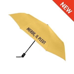 Puede incluir: Paraguas amarillo mostaza con la frase francesa "MERDE, IL PLEUT" en negro. El paraguas está abierto, mostrando su forma completa. Una etiqueta roja "NUEVO" está en la esquina superior derecha.