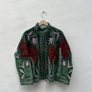 May include: A green velvet jacket with detailed floral embroidery in red and pink. The jacket has a high collar and is suspended on a wooden hanger. The embroidery provides a striking contrast against the dark green fabric.