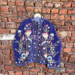 Könnte beinhalten: Blaue Samtjacke mit aufwendiger Blumenstickerei in Rosa, Weiß und Grün. Die Jacke hat einen Stehkragen und wird auf einem Kleiderbügel vor einer Backsteinmauer präsentiert. Der Text "NR TEXTILE" ist sichtbar.