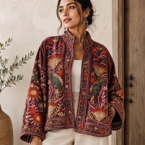 Pode incluir: Uma jaqueta bordô escuro com frente aberta, gola alta e mangas largas. A peça é decorada com intrincados bordados florais multicoloridos em tons de laranja, rosa, verde e dourado, combinada com uma regata branca e calças.