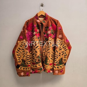 Könnte beinhalten: Eine braune Samtjacke mit lebendiger Stickerei von Tigern und Blumenmustern in Rosa, Grün und Gelb. Die Jacke hat einen Stehkragen, lange Ärmel und eine Vorderöffnung. Der Text NRTEXTILE ist auf der Vorderseite der Jacke deutlich zu sehen.