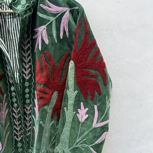May include: Green velvet jacket with embroidered floral design in red, pink, and green. The jacket features a striped lining and a collar with matching embroidery. A unique piece of apparel.