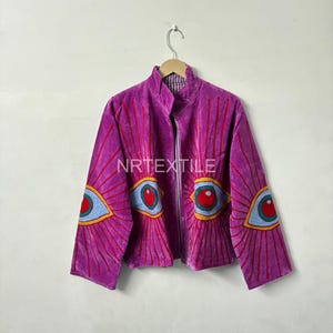 Könnte beinhalten: Eine leuchtend violette Jacke mit durchgehendem Reißverschluss, verziert mit gestickten Augenmotiven auf den Ärmeln und der Brust. Die Jacke hat einen hohen Kragen und hängt an einem Metallbügel. Der Text "NRTEXTILE" ist sichtbar.