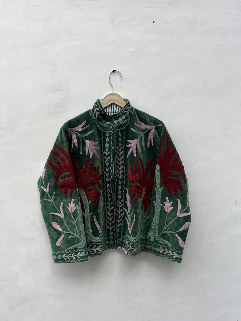 May include: A green velvet jacket with detailed floral embroidery in red and pink. The jacket has a high collar and is suspended on a wooden hanger. The embroidery provides a striking contrast against the dark green fabric.