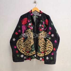Könnte beinhalten: Eine schwarze Jacke mit goldfarbenen, gestickten Tigern auf den Vorderteilen, umgeben von bunter Blumenstickerei in Rosa-, Rot- und Grüntönen. Die Jacke hat einen Stehkragen und ein schwarz-weiß gestreiftes Innenfutter. Der Text lautet NRTEXTILE.
