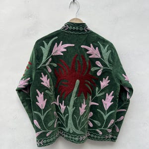 May include: A dark green velvet jacket with floral embroidery. The jacket features a large red flower design on the back, with pink and green leaves and stems. The jacket is hanging on a wooden hanger.
