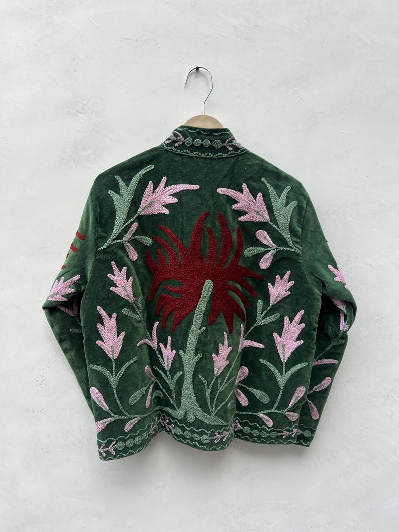 May include: A dark green velvet jacket with floral embroidery. The jacket features a large red flower design on the back, with pink and green leaves and stems. The jacket is hanging on a wooden hanger.
