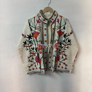 May include: Cream-colored embroidered jacket with a floral design. The jacket features a collared neckline and long sleeves. The embroidery includes orange, pink, teal, and black floral patterns. The jacket is hanging on a wooden hanger.