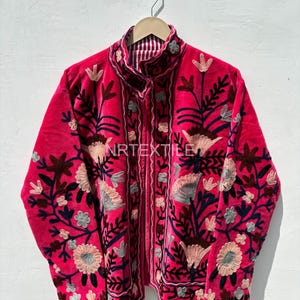 Könnte beinhalten: Eine leuchtend pinkfarbene Jacke mit aufwendiger Blumenstickerei in Braun-, Blau- und Cremetönen. Die Jacke hat einen hohen Kragen und ein gestreiftes Futter, was einen auffälligen Kontrast erzeugt. Das Design ist detailliert und auffällig.