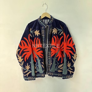 Könnte beinhalten: Eine marineblaue Jacke mit leuchtend orangefarbener Blumenstickerei. Die Jacke hat einen hohen Kragen und ein vertikales Streifenmuster in der Mitte. Der Text "NRTEXTILE" ist auf der Vorderseite zu sehen. Die Jacke hängt an einem Holzbügel.