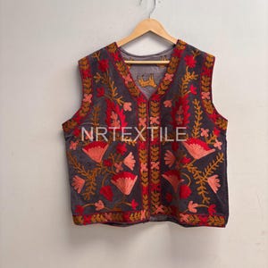 Könnte beinhalten: Eine ärmellose, dunkelgraue Weste mit aufwendiger Blumenstickerei in Rot-, Orange- und Goldtönen. Die Weste hängt an einem Holzbügel. Der Text "NRTEXTILE" ist unten sichtbar.