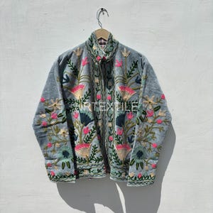 Könnte beinhalten: Hellblaue Jacke mit aufwendiger Blumenstickerei in Rosa, Grün und Gold. Die Jacke hat einen hohen Kragen und hängt an einem Holzbügel. Die Stickereidetails sind deutlich sichtbar.