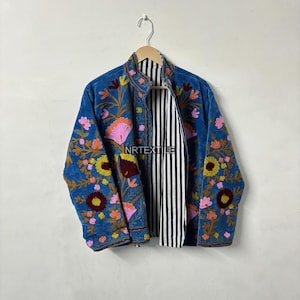 May include: A blue embroidered jacket with floral designs in pink, yellow, and orange. The jacket has a black and white striped lining and is hanging on a wooden hanger. The jacket is a medium weight and has a stand-up collar.