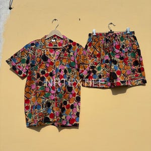 May include: A colorful pajama set featuring a short-sleeved button-down shirt and matching shorts. The set is adorned with a vibrant pattern of multicolored circles, outlined in white, on a beige background. The shirt has a chest pocket and white trim.