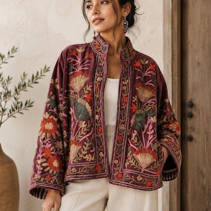 May include: A deep burgundy velvet jacket featuring a mandarin collar and wide sleeves. The garment is adorned with intricate, multi-coloured floral embroidery in shades of orange, green, and pink, covering the front panels and sleeve cuffs.