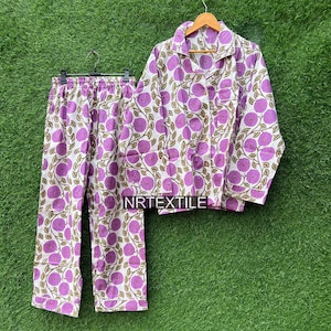 May include: A two-piece pajama set with a floral pattern. The set includes a button-down shirt and pants, both in white fabric with a repeating pattern of purple flowers and green leaves. The text "NRTEXTILE" is visible on the shirt.