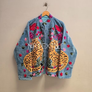 Könnte beinhalten: Eine hellblaue Jacke mit gestickten gelben Tigern mit schwarzen Streifen, umgeben von rosa und roten Blumenmotiven. Das Kleidungsstück hat einen Stehkragen, lange Ärmel und der Text "NPTEXTILE" ist auf der Vorderseite aufgedruckt.