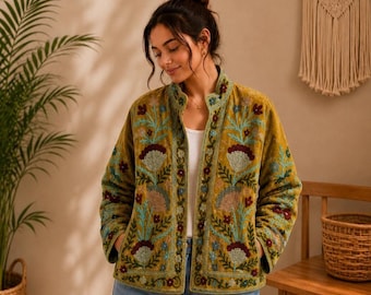 May include: A mustard yellow quilted jacket featuring a vibrant floral embroidery pattern in shades of teal, burgundy, and cream. The jacket has a high collar, long sleeves, and a straight hemline, worn over a plain white top and blue denim jeans.