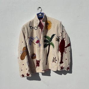 Könnte beinhalten: Beige Jacke mit gestickten Strandmotiven, darunter eine Sonne, eine Palme, ein Hummer und eine Sonnenbrille. Die Jacke hat ein rot-weiß gestreiftes Futter und hängt an einem Metallbügel.