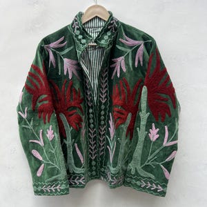 May include: A green velvet jacket with intricate floral embroidery in red and pink. The jacket features a collared neckline and a striped lining. The design includes detailed stitching along the edges and down the front, creating a unique and artistic garment.