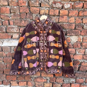 Könnte beinhalten: Braune Jacke mit gestickten Fischen in Orange-, Rosa- und Grüntönen. Die Jacke hat einen hohen Kragen und dekorative Stickereien am Kragen und an der Vorderseite. Die Jacke hängt an einem Holzbügel vor einer Backsteinmauer.
