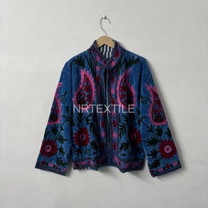 Könnte beinhalten: Blaue Samtjacke mit aufwendiger Blumenstickerei in Rosa und Rot. Die Jacke hat einen hohen Kragen und lange Ärmel. Der Text "NRTEXTILE" ist auf der Vorderseite der Jacke zu sehen.