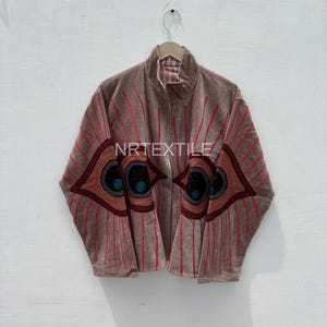 Könnte beinhalten: Braune Jacke mit einem einzigartigen Design. Die Jacke hat ein augenförmiges Design mit roten und braunen Akzenten. Der Text "NRTEXTILE" ist auf der Jacke sichtbar.