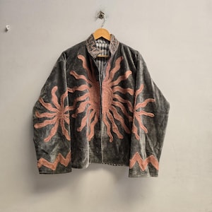 Puede incluir: Una chaqueta de terciopelo gris con motivos solares bordados en color melocotón en la parte delantera y las mangas. La chaqueta tiene cuello alto, mangas largas con patrones en zigzag en los puños y una abertura frontal, sobre una percha.