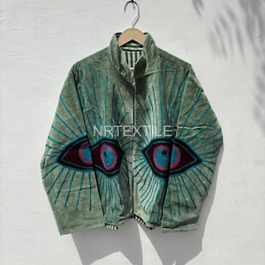 Könnte beinhalten: Smaragdgrüne Samtjacke mit gesticktem Augendesign. Die Jacke hat einen hohen Kragen und einen Reißverschluss. Die Augen sind mit türkis, weinrot und rosa Faden bestickt. Der Text "NRTEXTILE" ist sichtbar.