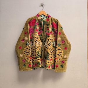 Könnte beinhalten: Eine beigefarbene Samtjacke mit lebendigen, mehrfarbigen Blumenstickereien und zwei großen, symmetrischen Tigermotiven auf den Vorderteilen. Das Kleidungsstück hat einen Stehkragen und der Text NRTEXTILE ist mittig auf der Brust aufgedruckt.