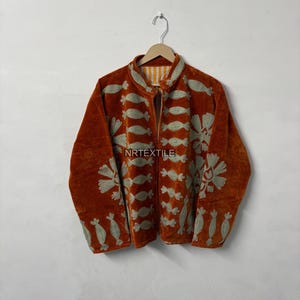 Könnte beinhalten: Rostorangefarbene Samtjacke mit aufwendigen Stickereien mit floralen und geometrischen Mustern. Die Jacke hat einen Stehkragen und einen durchgehenden Reißverschluss. Der Text "NRTEXTILE" ist auf der Vorderseite der Jacke zu sehen.