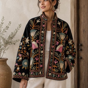 May include: A black open-front jacket featuring long sleeves and a mandarin collar, decorated with vibrant, multi-colored floral embroidery in shades of pink, blue, yellow, and green. The garment is worn over a white top and light-colored trousers.