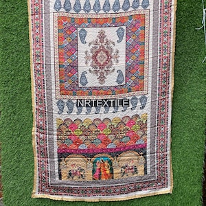 May include: A rectangular textile with a cream-colored background and intricate patterns. The design includes paisley shapes, geometric patterns, and a central floral motif. The edges are trimmed with gold-colored fabric, and the text "NRTEXTILE" is visible.