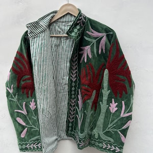 May include: A green velvet jacket with embroidered floral designs in red, pink, and green. The jacket has a striped lining and a collared neckline. The jacket is hanging on a wooden hanger.